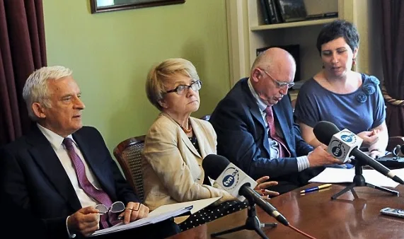 Panel of four people at a press conference seated at a table with microphones in front of them, one speaking into a recorder while others listen and take notes.