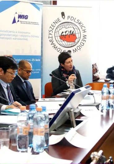 Panel discussion at a conference: a woman speaks into a microphone at a long table, flanked by two men, with banners and water bottles in the background.
