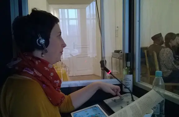 Female radio host wearing a headset and orange scarf speaks into a microphone in a recording studio, looking at notes.