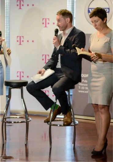 Man in a dark suit sits on a bar stool on stage, speaking into a microphone while a woman beside him reads from a book; T?Mobile logos on the backdrop.