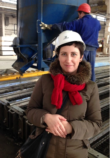 Woman in a white hard hat, red scarf, and brown coat at an industrial site with large blue machinery behind her; a worker in a red helmet is in the background.