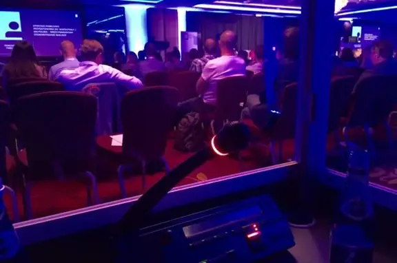 Audience seated in a dimly lit conference room with purple lighting, watching a presentation on stage with screens at the front.